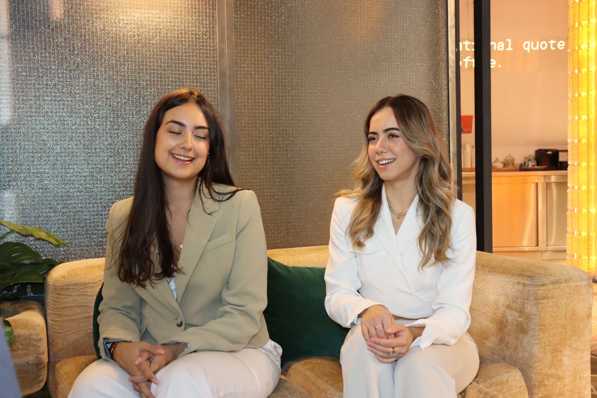 Two women in blazers sit side by side on a golden sofa, smiling as they listen